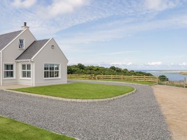 A house with white walls and gray roof by a gravel driveway and green lawn near a wooden fence with water and bushes in the background at The Haven in Falcarragh County Donegal