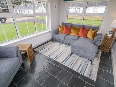 A sitting area with a gray sectional sofa with orange and yellow pillows a wooden side table and a lamp on a wooden stand on a rug over black tiled floor with large windows showing outside at The Haven in Falcarragh County Donegal