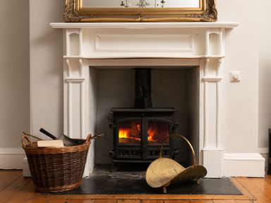 A wood burning stove in a white fireplace with a basket of firewood on the left and a brass firewood holder on the right at Fron Dderw in Bala