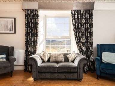 A living room with a gray sofa and patterned cushions between two chairs by a window with floral curtains at Fron Dderw in Bala