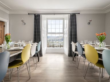 A dining room with tables, colorful chairs, glassware, plates, tulip flowers, and a window with dark curtains at Fron Dderw Bala