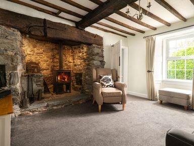 A living room with a stone fireplace and wood-burning stove an armchair with a cushion a window with curtains and a footstool at Fron Dderw in Bala