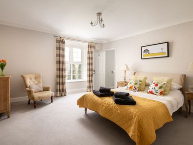 A bedroom with a double bed with yellow quilt and floral pillows wooden furniture an armchair and striped curtains at Fron Dderw in Bala