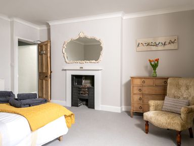 A bedroom with a bed covered in yellow quilt and gray towels wooden door a fireplace with a decorative mirror above a wooden chest of drawers with flowers and an upholstered armchair at Fron Dderw in Bala