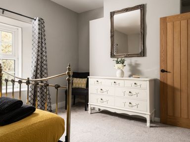 A bedroom with a metal bed frame yellow bedding folded towels a white dresser with a vase and mirror a chair near a window with patterned curtains at Fron Dderw in Bala