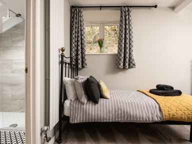 A bedroom with a single bed with striped bedding and pillows by a window with patterned curtains and a vase of flowers with an adjacent bathroom at Fron Dderw in Bala