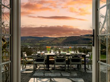 An outdoor dining table set with glassware and vases of flowers on a stone patio overlooking hills and a lake at Fron Dderw Bala