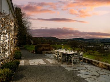 An outdoor dining table set with chairs and flowers on a stone patio overlooking a green landscape at Fron Dderw in Bala