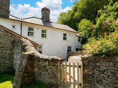 A white stone house with a slate roof behind a stone wall and wooden gate with greenery around at Fron Dderw Bala