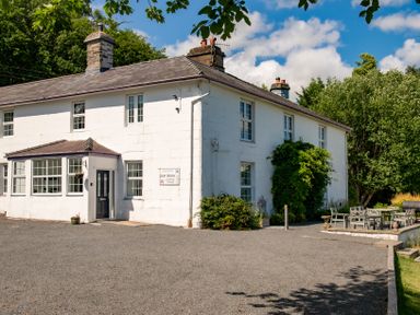 The exterior of a white two-story house with multiple windows and a gravel driveway outdoor seating area with tables and chairs surrounded by trees and greenery at Fron Dderw in Bala
