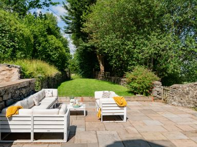 An outdoor patio area with white cushioned seating and a coffee table surrounded by stone walls and greenery at Fron Dderw in Bala