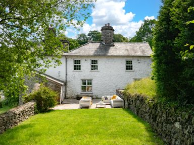 An outdoor patio with white seating and tables behind a white stone house with green grass and trees at Fron Dderw in Bala