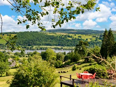 A view of a grassy hill with trees and a river in the background and a wooden deck with red chairs and a table at Fron Dderw in Bala