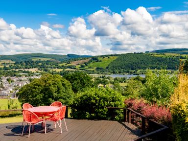 A deck with a red table and four chairs overlooking a valley with trees hills and a body of water at Fron Dderw in Bala