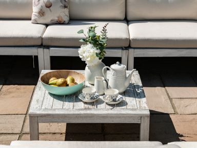 A wooden table with a bowl of pears white teapot cups and a vase with flowers on a patio with cushioned seating at Fron Dderw Bala