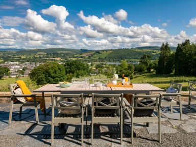 An outdoor dining table with chairs on a stone terrace overlooking hills and a village at Fron Dderw in Bala