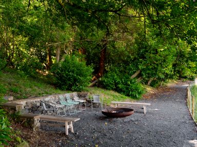An outdoor seating area with metal chairs wooden benches and a fire pit surrounded by trees and a gravel path at Fron Dderw in Bala