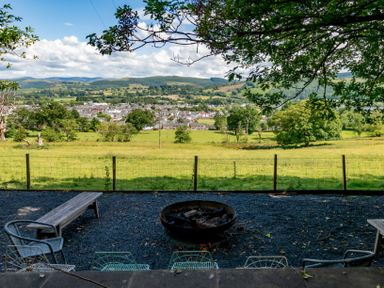 An outdoor seating area with metal chairs and wooden benches around a fire pit overlooking a grassy field and a town with hills in the background at Fron Dderw in Bala