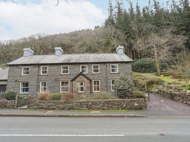 A stone cottage with multiple chimneys and windows by a road near a wooded hill at Glyn Awel in Nant Gwynant near Beddgelert