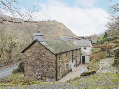 Stone cottages with chimneys near a road and hillside with trees at Glyn Awel in Nant Gwynant near Beddgelert