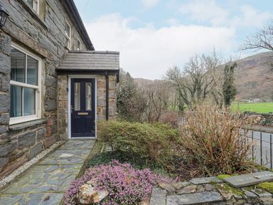 An entrance door to a stone house with a stone path and garden with bushes and flowers at Glyn Awel in Nant Gwynant near Beddgelert