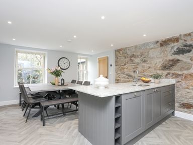 A kitchen with a gray island, stone wall, dining table with chairs and bench with flowers at Glyn Awel in Nant Gwynant near Beddgelert