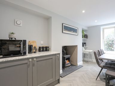 A kitchen area with grey cabinets microwave kettle and canisters next to a wood stove and dining table with chairs at Glyn Awel in Nant Gwynant near Beddgelert