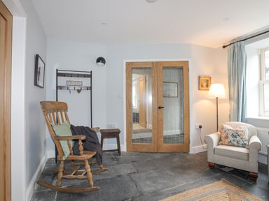 A sitting area with a wooden rocking chair with cushions and a throw a small wooden stool a cushioned armchair with a floral pillow a floor lamp a window with curtains wooden framed glass doors and a coat rack with a croeso sign at Glyn Awel in Nant Gwynant near Beddgelert