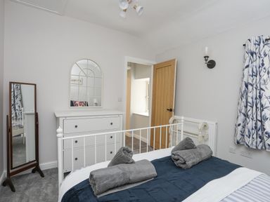 A bedroom with a white metal bed frame blue bedding gray towels a white dresser a standing mirror an open wooden door and patterned curtains at Glyn Awel Nant Gwynant near Beddgelert