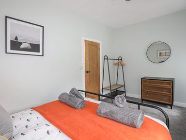 A bedroom with a bed covered with an orange blanket and gray towels a wooden door a clothing rack a dresser with three drawers and a round mirror on the wall at Glyn Awel in Nant Gwynant near Beddgelert