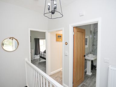 A hallway with white walls two open doors revealing a bedroom and a bathroom a round mirror hanging on the wall and a light fixture hanging from the ceiling at Glyn Awel in Nant Gwynant near Beddgelert