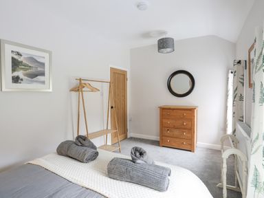 A bedroom with a bed holding rolled towels a wooden chest of drawers a round mirror a clothes rack and floral curtains at Glyn Awel in Nant Gwynant near Beddgelert