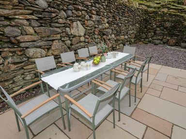 Outdoor patio area with a glass table and eight chairs against a stone wall at Glyn Awel in Nant Gwynant near Beddgelert