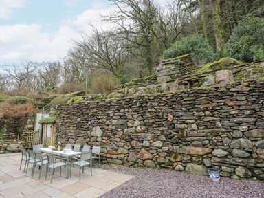 An outdoor dining table with chairs on a stone patio next to a stone retaining wall with trees beyond at Glyn Awel in Nant Gwynant near Beddgelert