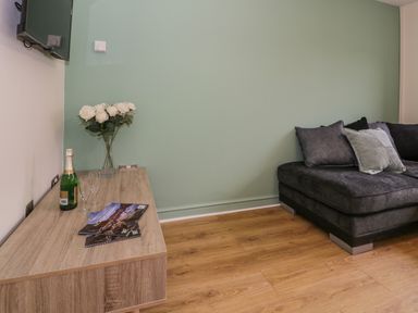A corner of a living room with a wooden cabinet holding a vase of white flowers a bottle of champagne two glasses and magazines next to a dark gray sofa at Bwythyn Yr Wyddfa in Llanberis