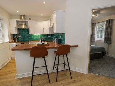 A kitchen with a wooden breakfast bar with two brown stools a teal tile backsplash and white cabinets at Bwythyn Yr Wyddfa in Llanberis