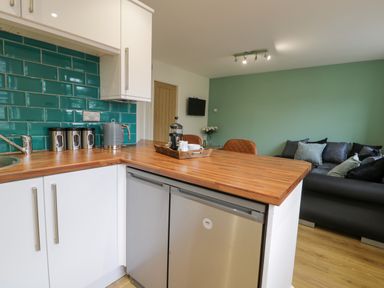 A kitchen corner with wooden countertops white cabinets teal subway tile backsplash and a living area with a sofa and green wall at Bwythyn Yr Wyddfa in Llanberis