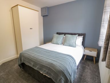 A bedroom with a double bed, two cushions, a blue throw, a white wardrobe, a wooden nightstand, and curtains at Bwythyn Yr Wyddfa in Llanberis