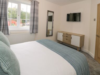 A bedroom with a bed, wall-mounted TV, wall mirror, window with curtains, and a wooden cabinet at Bwythyn Yr Wyddfa in Llanberis