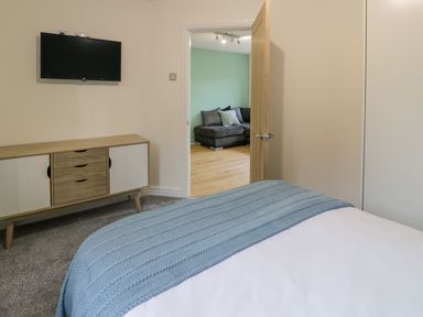 A bedroom with a blue blanket on the bed a wooden cabinet and a wall-mounted TV looking into a living room with a gray sofa at Bwythyn Yr Wyddfa in Llanberis