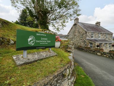 A stone house near a green welcome sign for Lyons Snowdon View Holiday Park with grass and a tree in the background