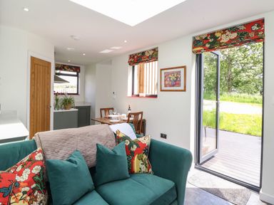A living area with green sofa and floral cushions next to a dining table and open door to garden at Brow Wood Cabin in Ullswater