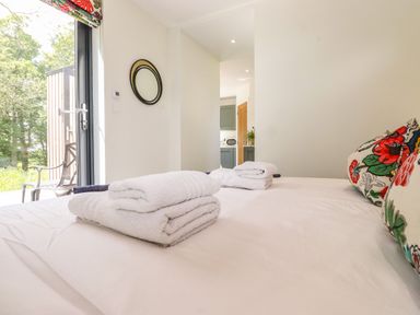 A bedroom with a bed holding folded white towels and floral pillows with a view of a kitchen and glass door to outside at Brow Wood Cabin Ullswater