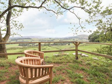 Two wooden chairs on a grass area near a wooden railing overlooking fields and hills at Brow Wood Cabin in Ullswater