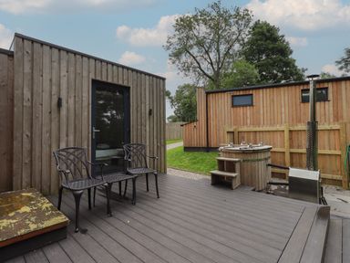An outdoor patio with two black chairs a wooden hot tub and a wooden cabin in the background at Brow Wood Cabin Ullswater