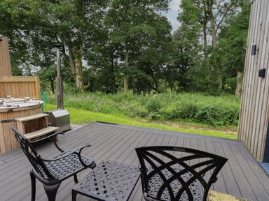 An outdoor deck with two black metal chairs a small matching table a wooden hot tub and green trees in the background at Brow Wood Cabin in Ullswater