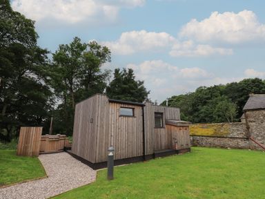A small wooden cabin with a gravel path and a wooden hot tub near a stone wall at Brow Wood Cabin in Ullswater