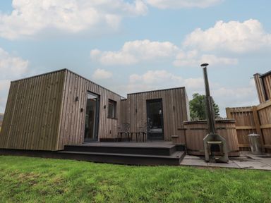 A wooden cabin with a small deck and two chairs next to a hot tub and outdoor stove at Brow Wood Cabin in Ullswater