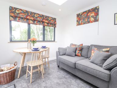 A living room with a grey sofa, wooden chairs, a round wooden table with dishes and flowers and floral curtains at Bluebell Cabin in Ullswater