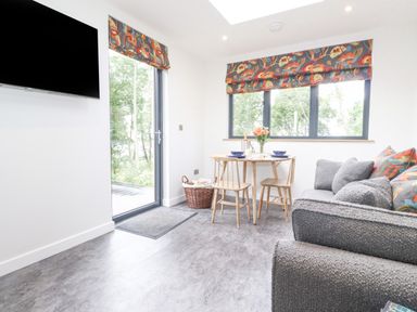 A sitting area with a gray sofa a small wooden dining table with two chairs and windows with floral blinds at Bluebell Cabin in Ullswater